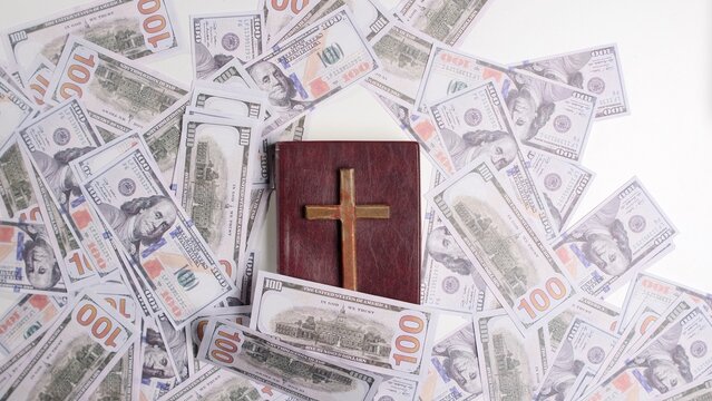 Christian bible with a golden cross surrounded by numerous hundred dollar bills, illustrating concepts of religion, finance, tithing, wealth, and spiritual donations