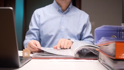 Person is reviewing a binder full of documents on an office desk, with laptops and a small wooden...