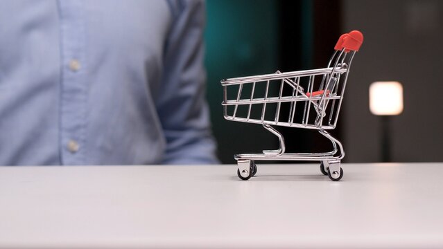 Miniature shopping cart standing on a white table with an unidentifiable person in the background, symbolizing e commerce, shopping, and retail business concepts
