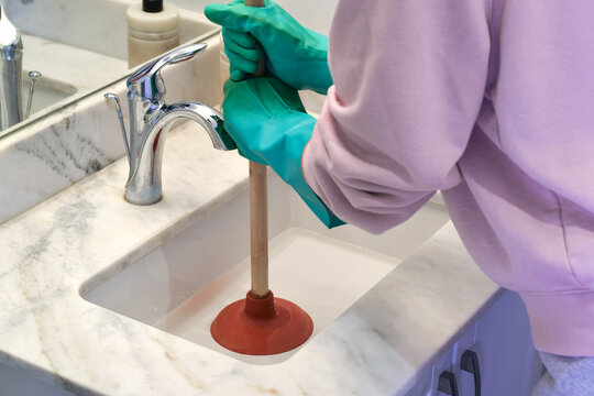 Woman unclogging bathroom sink drain with plunger at home. Everyday household maintenance concept showing plumbing problem, blockage removal, cleaning, and domestic repair routine
