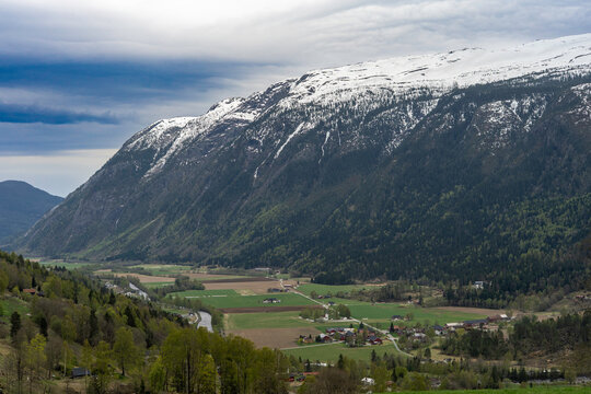A breathtaking view of Norway's mountainous landscape. Snow-capped mountain ranges frame a beautiful valley with village houses, fields, and a river. Cloudy spring day.  Norwegian scenery traveling.