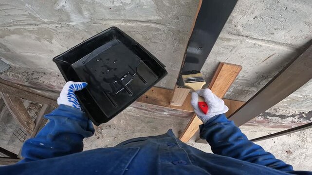 The process of painting wood. A builder paints a wooden beam black. A worker in a blue uniform and protective gloves applies paint with a brush. First-person view (POV). Redesign, handmade