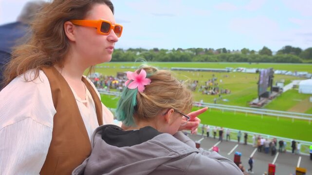 A girl and a woman look from the grandstand of a hippodrome at the crowd of spectators waiting for the horse race. Good mood. Day off. Family leisure outdoor event