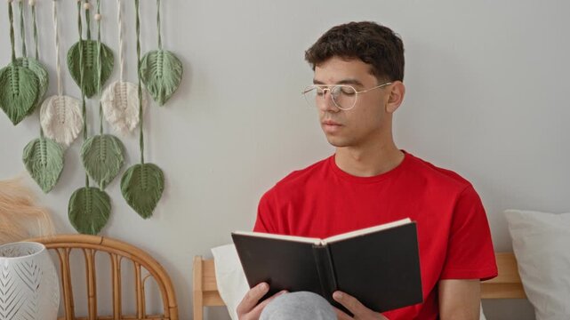 Man reading book on bed inside building, holding the hardcover with both hands while seated and wearing glasses; quiet contemplation calm.