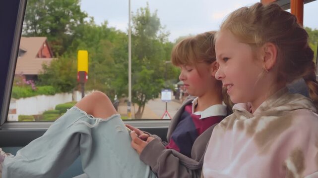 Two smiling girls ride on the upper deck of a city bus. They look out the window and smile at each other. Views of Liverpool suburbs outside. Happy mood
