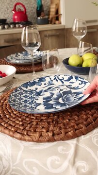 Table setting video montage with elegant home dining atmosphere. Hands arranging blue floral plates and glassware for stylish tablescape. Cozy kitchen scene with decorative woven placemats and warm