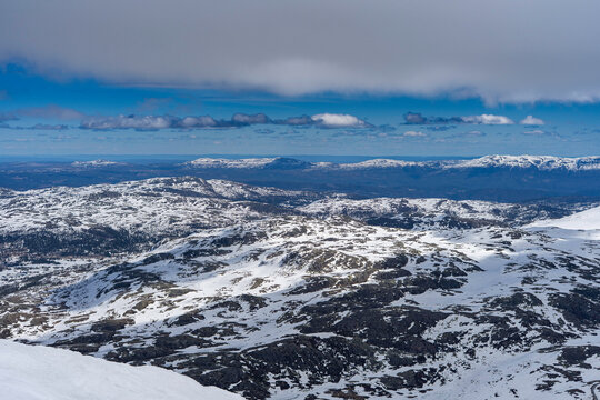 Panoramic view from the summit of Gaustatoppen at 1,850 meters above sea level. Snow-covered mountain plateaus, rocky ridges, and vast landscapes of southern Norway under a dramatic cloudy sky.
