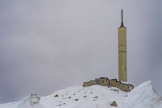 Winter view of the iconic communication tower and mountain facilities on the summit of Gaustatoppen. Snow-covered rocky terrain under a dramatic overcast sky in the Norwegian mountains. 
