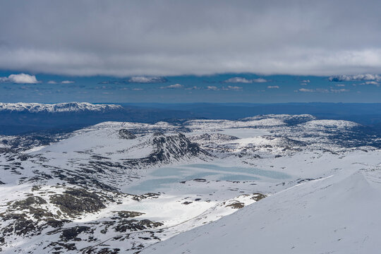 Panoramic view from the summit of Gaustatoppen at 1,850 meters above sea level. Snow-covered mountain plateaus, rocky ridges, and vast landscapes of southern Norway under a dramatic cloudy sky.
