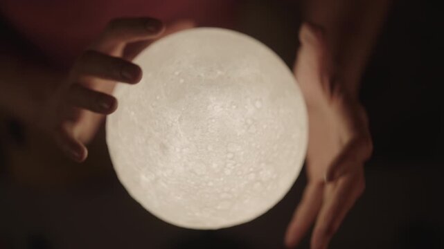 Closeup of fortune teller divining future using glowing crystal moon ball during mystical session, hands moving with magical energy over mysterious lunar sphere at night in dark interior, slow motion.