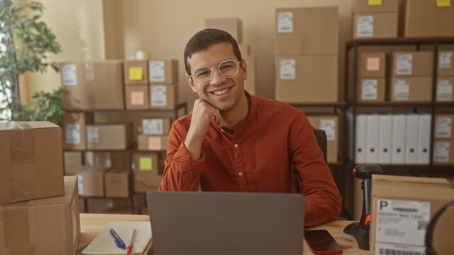 Man hand on chin smiling at laptop amid stacked shipping boxes in a building; small business optimism.