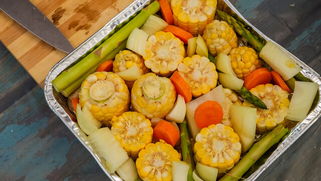 Freshly prepared corn, carrots, and asparagus arranged in a foil tray on a rustic wooden surface with a knife nearby
