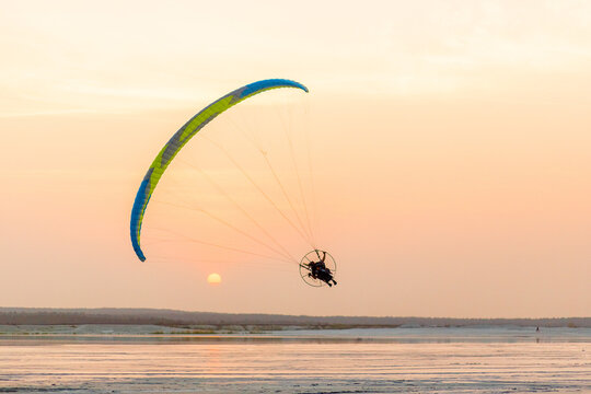 A motorized paraglider soars over a calm water surface, with a vibrant green and blue canopy catching the eye against the soft sky backdrop