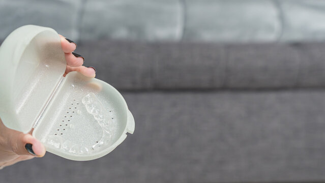 Hand holding a transparent case with a clear dental aligner inside, set against a blurred gray sofa background
