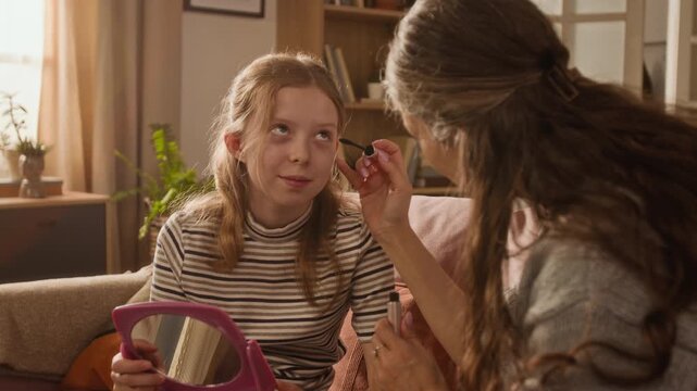 Medium shot of adult mother applying mascara to lashes of girl child while teaching daughter how to make up using cosmetics in living room