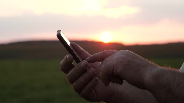 Close up of man hands holding and using smartphone apps. Scrolling through screen with finger. Against golden sunset over green field outdoor. Person using messenger to writing a message to relatives