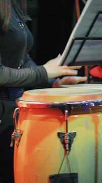 A female musician plays conga drums on stage during a live jazz performance. This close-up shot focuses on the rhythmic action, with warm, dramatic lighting highlighting the instrument and hands