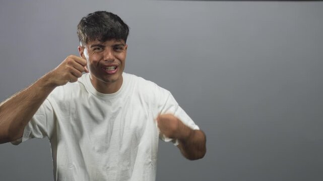 Young hispanic man tugs cheeks, shapes an l with hand and sticks out tongue, showing face and hand gestures in studio gray backdrop; playful mischief.
