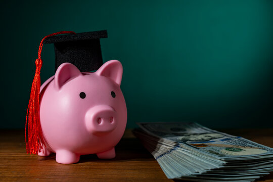 Pink piggy bank wearing a graduation mortarboard hat next to stack of american dollar banknotes. Concept of saving for college education cost student loans and financial planning