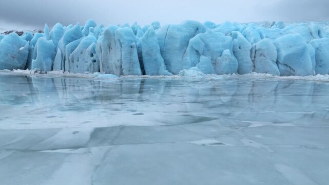 Aerial view of the Jokulsarlon glacier lagoon featuring massive blue ice formations reflecting on the frozen water surface under a cloudy sky in Sveitarfelagid Hornafjordur, Iceland.