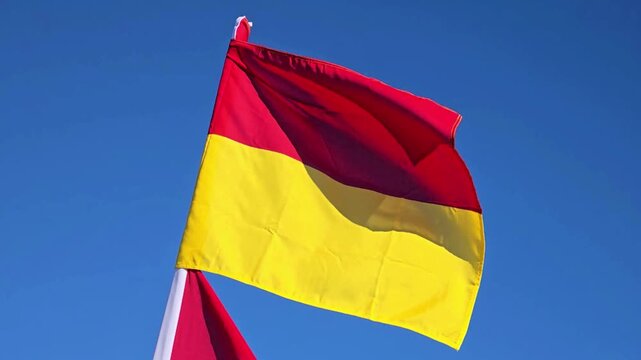 Red and yellow warning flag waving against a clear blue sky on a sunny day.