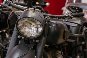 Fototapeta premium Detailed close-up view of vintage motorcycle front, showcasing large round glass headlight with ribbed texture and matte black housing. Handlebars and part of fuel tank are visible against a blurred