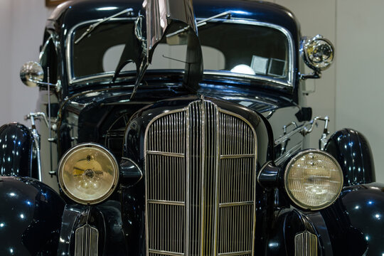 Detailed front view of classic dark blue automobile showcasing chrome grille, circular headlights, and partially open hood. Indoor setting highlights the car's glossy finish and elegant design.