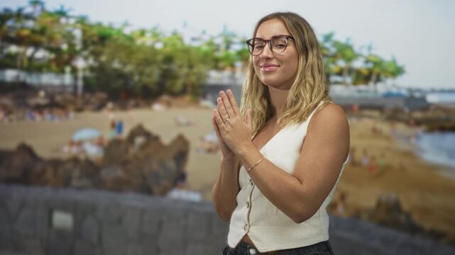 Young blonde woman wearing glasses and a sleeveless vest, hands clasped at chest in a prayerlike gesture on a sandy beach with palm trees and people visible; serenity.