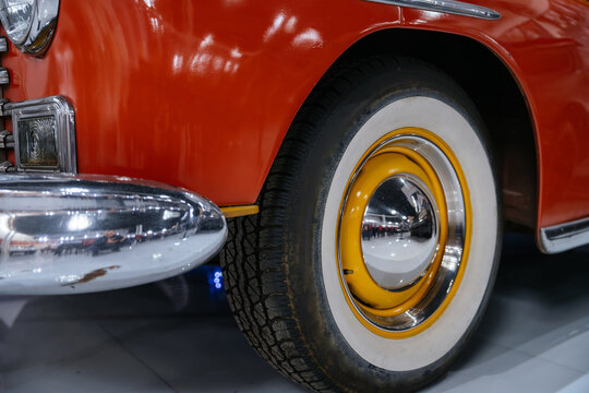 Detailed close-up captures front wheel and bumper of vintage orange automobile. Features white sidewall tire, yellow rim, and chrome hubcap reflecting showroom interior.