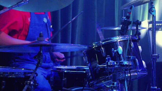 Close up of drummer in red shirt and blue overalls playing drum kit. Dynamic scene with motion blur on sticks and hands under green stage lighting. Energetic live music performance concept
