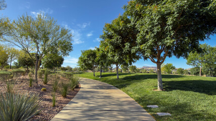 Obraz premium Vibrant view of lush green hills and manicured lawns in a public park. Featuring winding walking paths, decorative shrubs, and leafy trees under a clear blue sky. Peaceful summer day atmosphere.