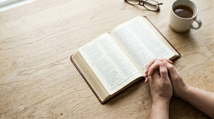 Naklejka premium Faith and prayer with open Bible on table beside cup of coffee, hands clasped in devotion. Faith in Christianity promotes reflection and connection,
