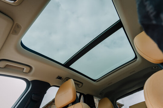 Interior view of a modern car with a panoramic sunroof showcasing a cloudy sky and beige leather seats