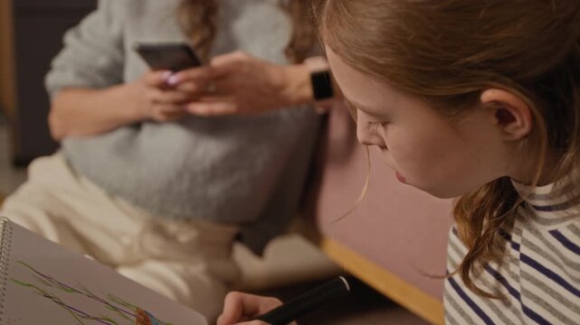 Tilt up shot of young girl drawing sketches with felt tip pen spending time with mother while woman messaging on smartphone distracted by online content