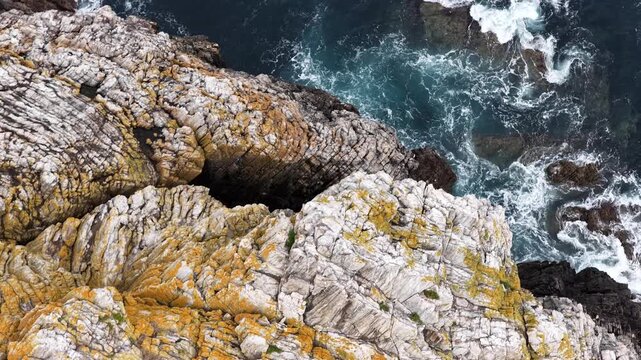 Aerial view of Silence Beach featuring rugged rock formations with yellow lichen and dark blue ocean waves crashing against the shore in Asturias, Asturias, Spain.