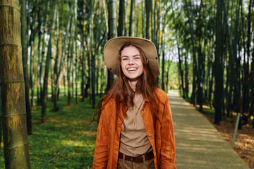 Fototapeta premium Woman smile hat bamboo forest path, portrait of a happy young female walking outdoors on a sunlit boardwalk, casual jacket and straw hat, cheerful candid lifestyle shot in nature.