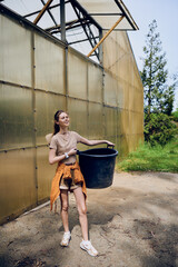 Fototapeta premium Woman holding bucket near greenhouse, smiling in outdoor setting while gardening. Young gardener in casual outfit with tied sweater and sneakers, carrying large plastic container on sunny day.