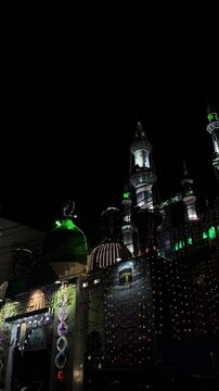 Night view of a mosque exterior featuring a green dome and white minarets illuminated by colorful string lights against a dark sky during 12 Rabi ul Awal (Eid Milad un Nabi) in Pakistan.