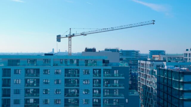 Tower crane rotating over modern city residential area. Tall construction tower crane rotating its jib over new residential buildings against a clear blue sky, showing urban development