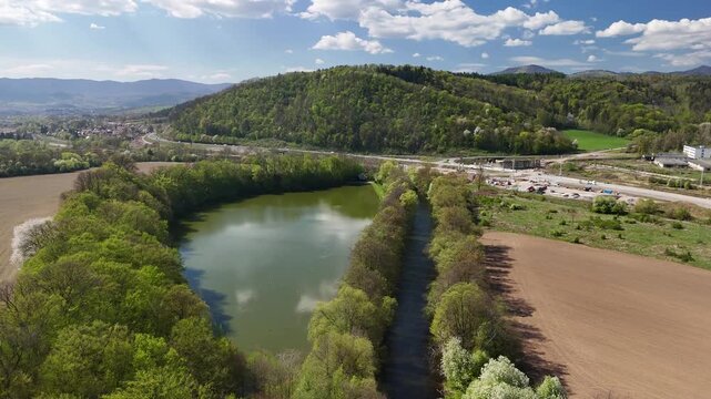 Aerial view of Salkova pond surrounded by lush green trees and fields with a forested hill under a blue sky in Banska Bystrica, Banskobystricky kraj, Slovakia.