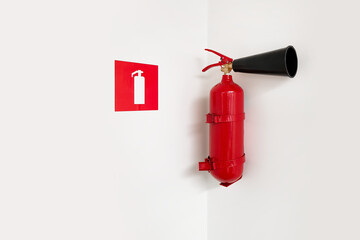 Naklejka premium A red fire extinguisher mounted in the corner of a white wall next to a fire safety sign. Minimalist image emphasizing industrial safety and precautionary measures in office or public spaces.