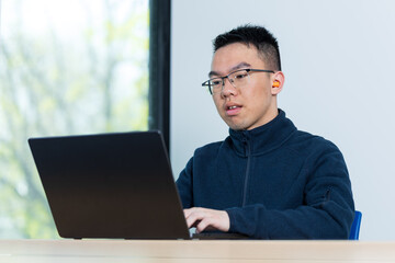 A focused Asian male student wearing glasses and orange earplugs types on a laptop at a desk by a window, ideal for neurodiversity, sensory sensitivity, or distraction free study concepts. © fotolite