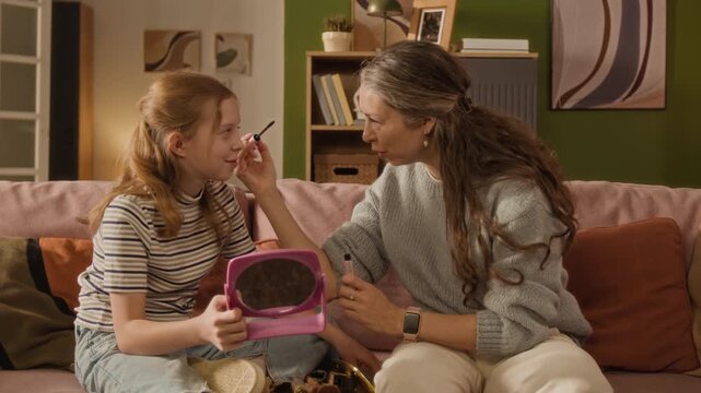 Side view of middle aged woman applying mascara to lashes of preteen daughter while putting on morning makeup together sitting on couch in living room
