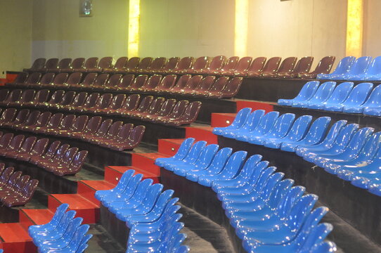 Wide shot of empty indoor stadium with blue and brown seats arranged in tiers, intersected by red stairs and warm lighting columns, showcasing architectural symmetry and vibrant color contrast