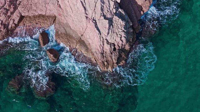 Top down drone shot of waves crashing against rocky coastline with turquoise water. Cinematic aerial movement ideal for nature, texture, and coastal concepts.