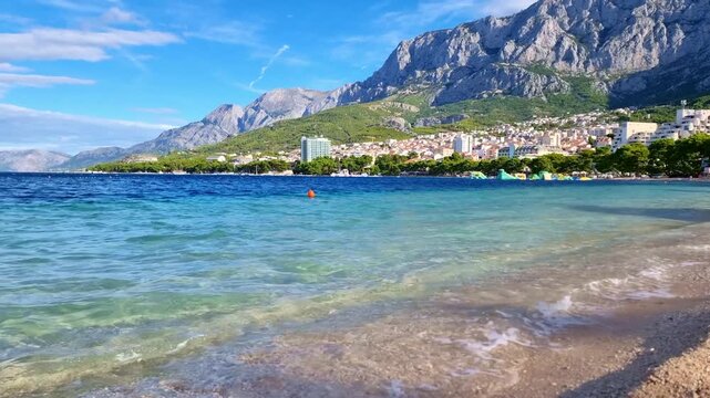 The turquoise water of the Adriatic Sea breaking on the Makarska town main pebble beach and the Biokovo mountain, Croatia, in sunny summer day
