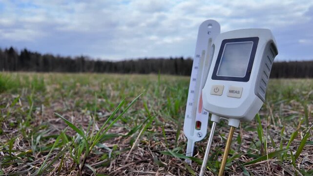 close-up, ground-level view of grass where a soil thermometer is already inserted into the earth. A hand places a pH meter into the soil for measurement. Scene represents agricultural testing