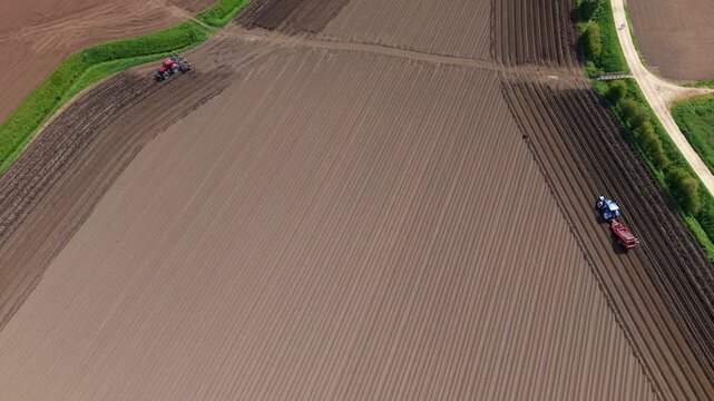 Aerial view of two tractors plowing and tilling a large brown agricultural field with neat soil rows and green borders in Knottingley, England, United Kingdom.