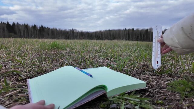 Ground-level close-up of an open notebook on soil while a hand inserts a soil thermometer followed by a pH meter into the ground. The device is turned on and the display lights up, after which data is