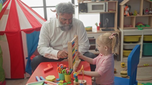 Man guides toddler moving abacus beads at red table in building playroom, pointing to rows and teaching counting; tenderness learning bond.